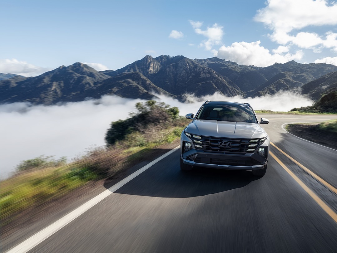 Gray Hyundai Tucson driving on a mountain road with blue sky, white clouds, and mountain range background, showcasing automotive design.