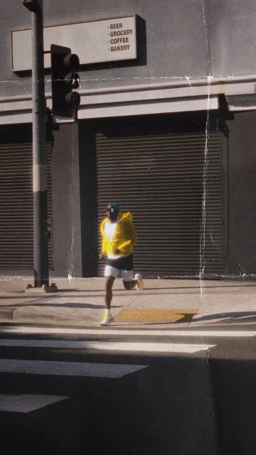 Person in a yellow jacket runs across a crosswalk in an urban setting near a store with a sign that reads 'Beer, Grocery, Coffee, Bakery'.