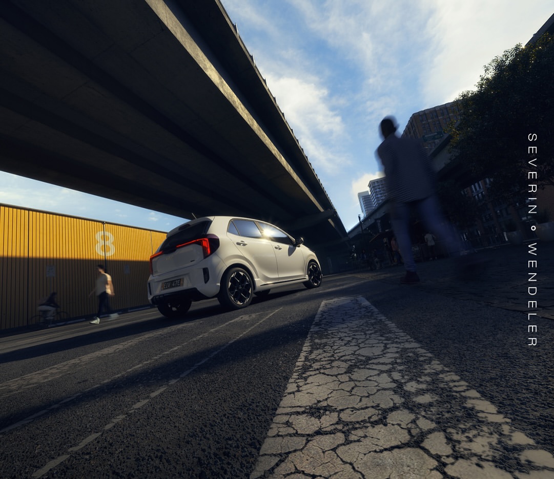 A white compact car drives on a city street under a bridge on a sunny day, with pedestrians walking by and buildings in the background.