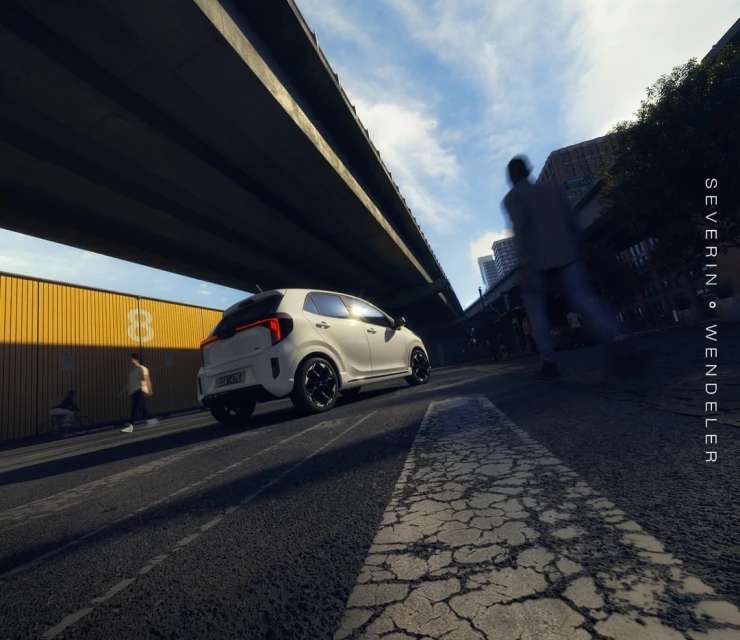 A white compact car drives on a city street under a bridge on a sunny day, with pedestrians walking by and buildings in the background.