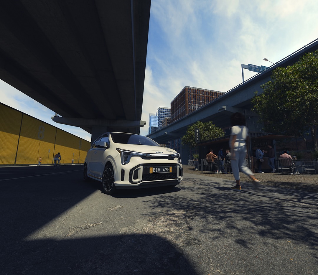 White Kia Niro EV under an overpass with pedestrians nearby. The compact electric car is parked on asphalt in an urban environment.
