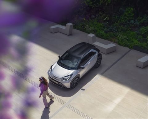 A silver compact car parked on a concrete surface with a woman walking by. Landscaping and architectural benches are in the background.