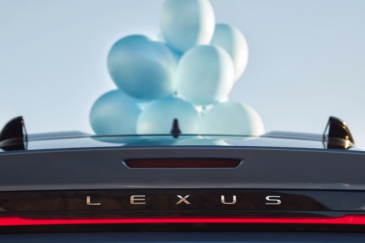 Rear view of a dark-grey Lexus car with blue balloons on the roof and its name written on the trunk against a light-blue sky.