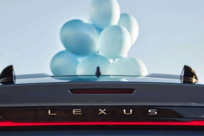 Rear view of a dark-grey Lexus car with blue balloons on the roof and its name written on the trunk against a light-blue sky.