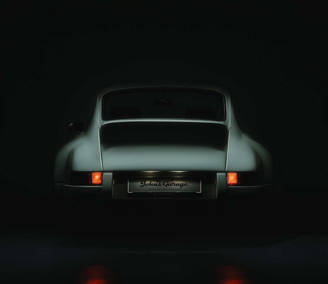 Rear view of a classic silver sports car in a dark setting, lights reflecting off the glossy floor, with a 'Johns Garage' license plate.