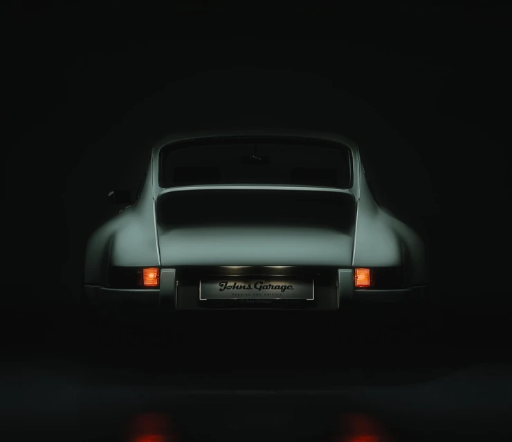 Rear view of a classic silver sports car in a dark setting, lights reflecting off the glossy floor, with a 'Johns Garage' license plate.