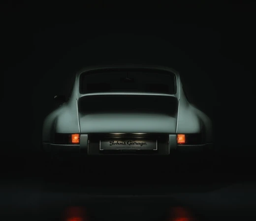 Rear view of a classic silver sports car in a dark setting, lights reflecting off the glossy floor, with a 'Johns Garage' license plate.