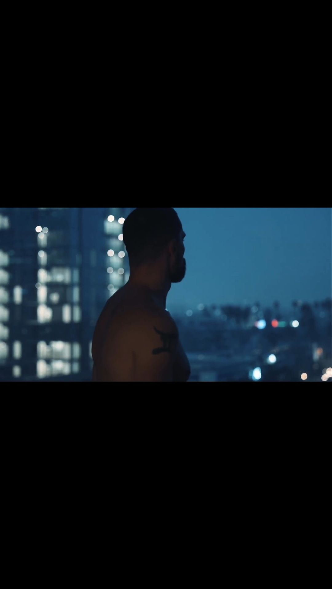 Man stands on a balcony overlooking a nighttime cityscape with illuminated buildings under a cloudy sky, creating a contemplative mood.