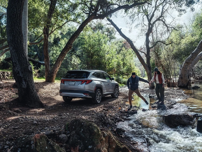 Silver SUV parked near a wooded area with a couple holding hands and crossing a rocky stream on a sunny day in a natural outdoor setting.