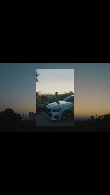 A stylish male model poses on the hood of a white BMW with a cityscape view at dusk, showcasing automotive elegance and urban lifestyle.
