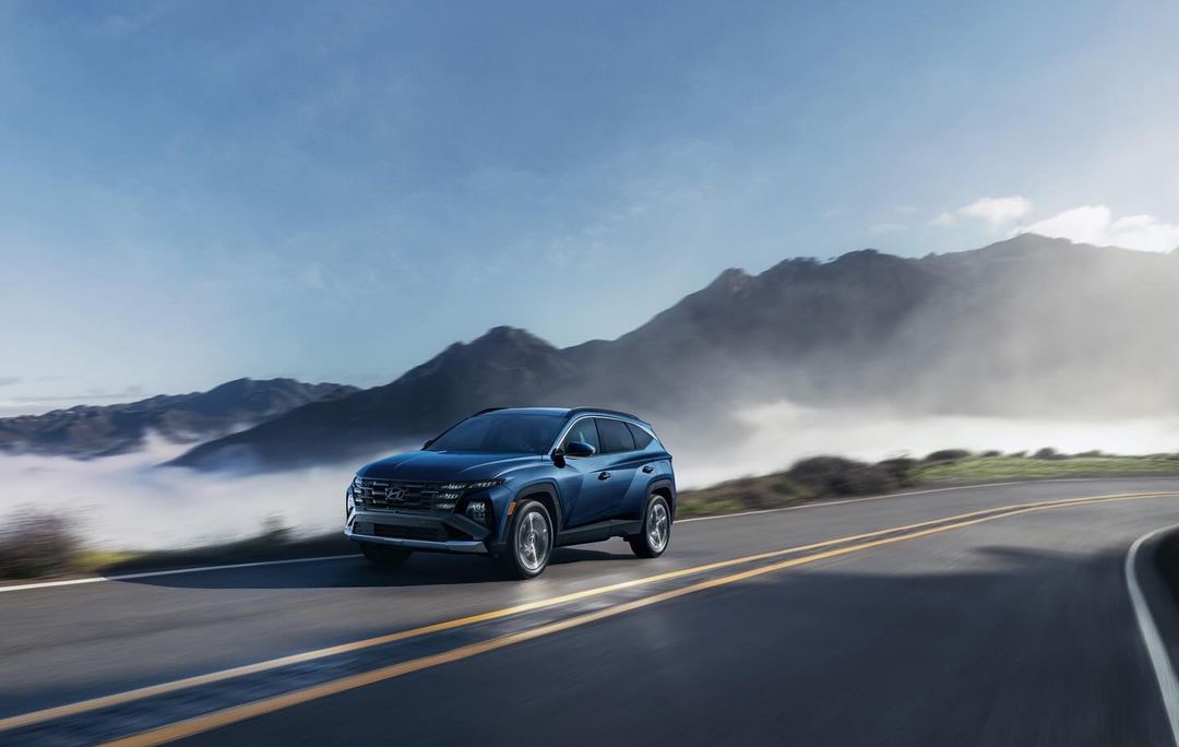 Blue Hyundai Tucson SUV driving on an asphalt road with mountains and fog in the background on a sunny day, automotive photography.