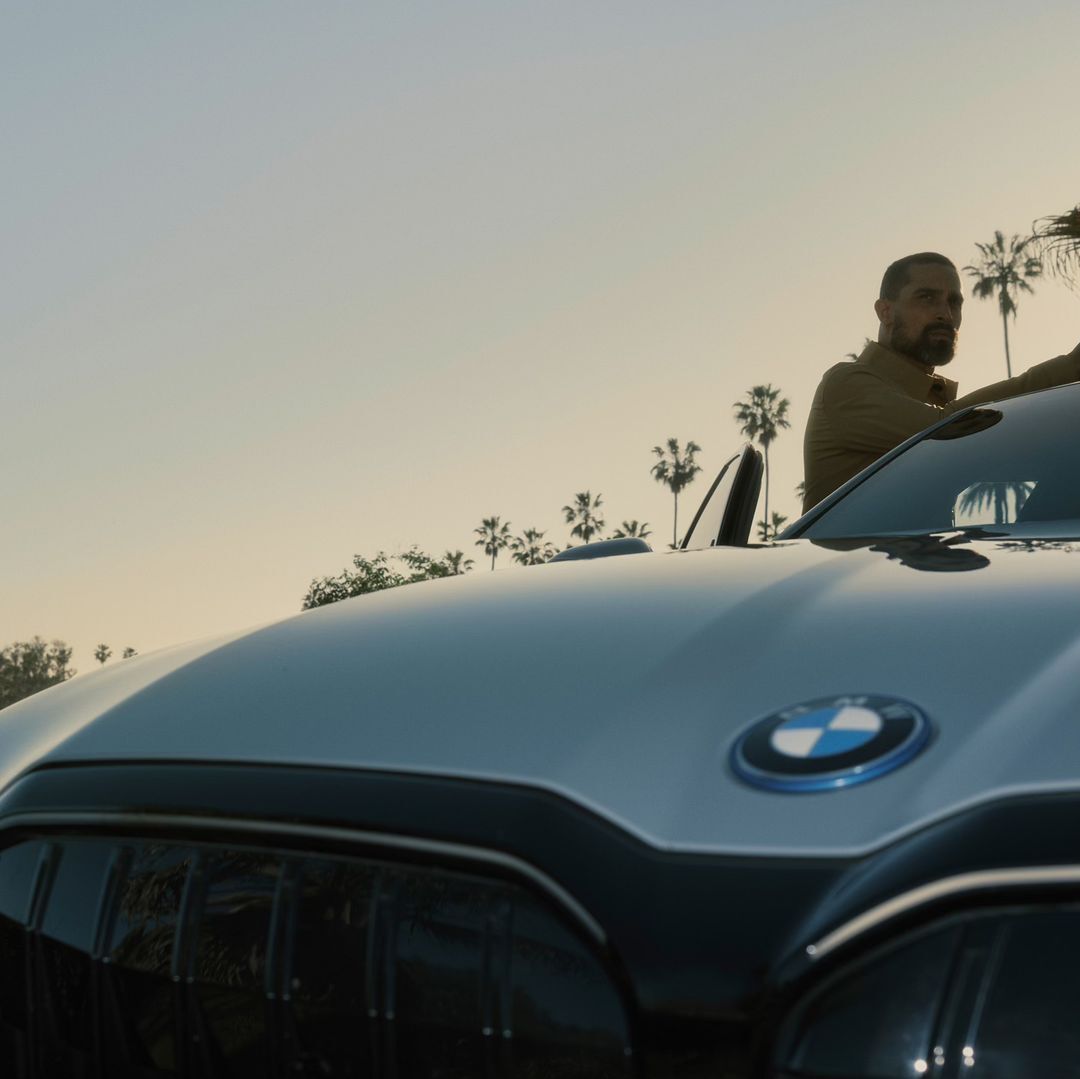 Silver BMW parked outdoors in a sunny, tree-lined setting, showcasing its front design with a bearded adult male visible in the background.