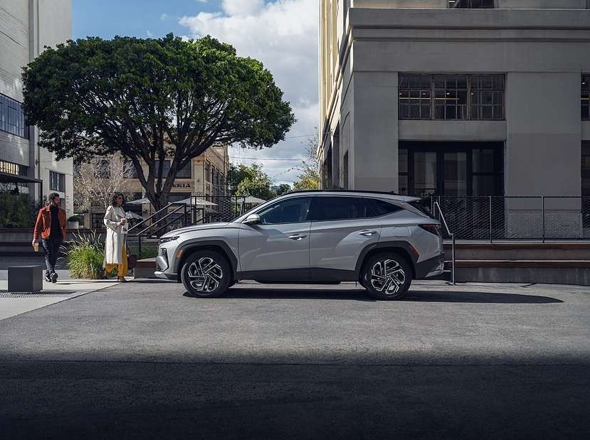 Silver SUV parked on an asphalt city street near pedestrians, buildings, and a green leafy tree under a blue sky with clouds.