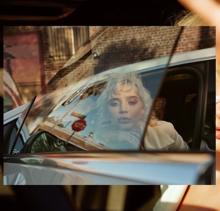 Woman with platinum blonde hair looks out from a car window in front of a brick building with a stop sign visible in the background.