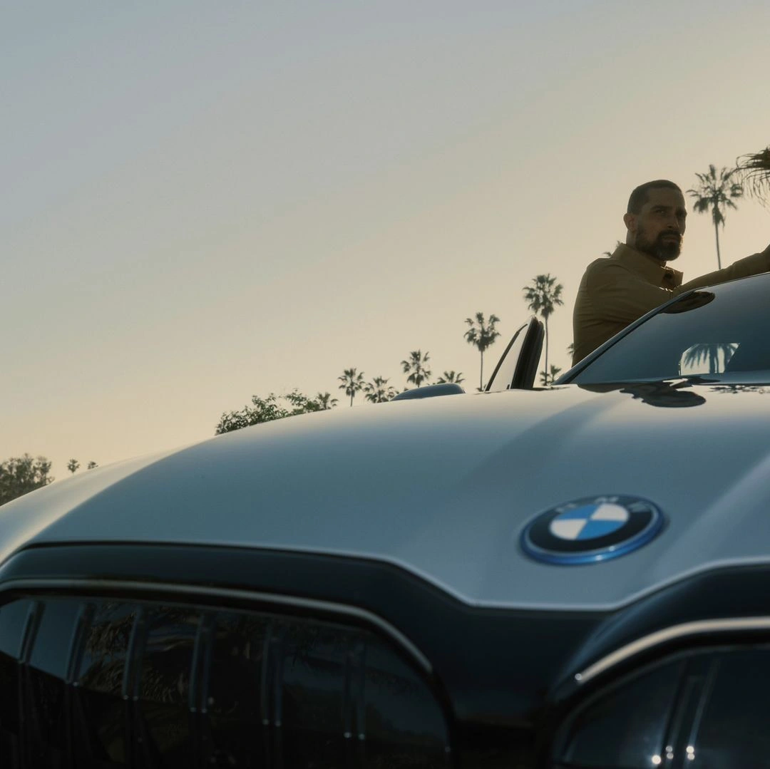 Man standing next to a luxury car outdoors with palm trees under a bright sky, captured by photographers Dejan and Per.