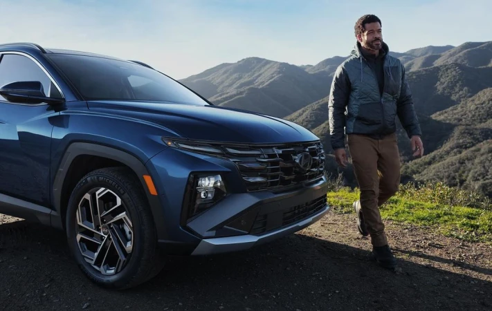 A man standing next to a blue SUV parked on a dirt path with mountain ridges in the background.
