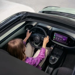 High-angle view of a woman driving a compact car with her hands on the steering wheel, wearing a purple jacket.