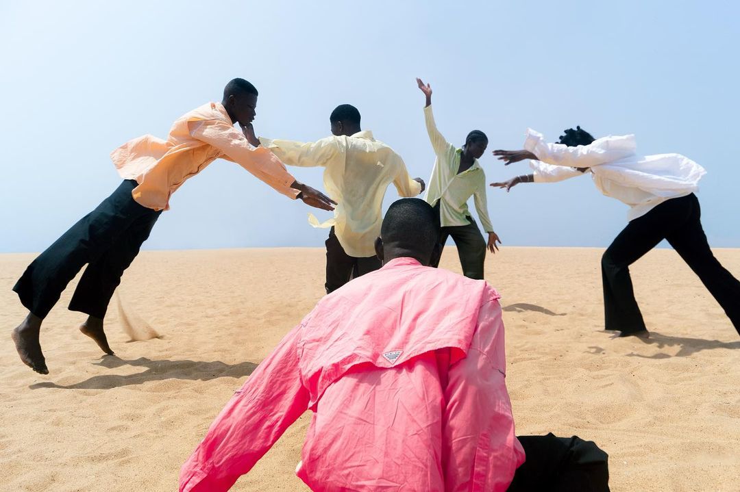 Group of young African men in shirts and black pants performing barefoot on a tan sand dune against a clear blue sky in bright daylight.