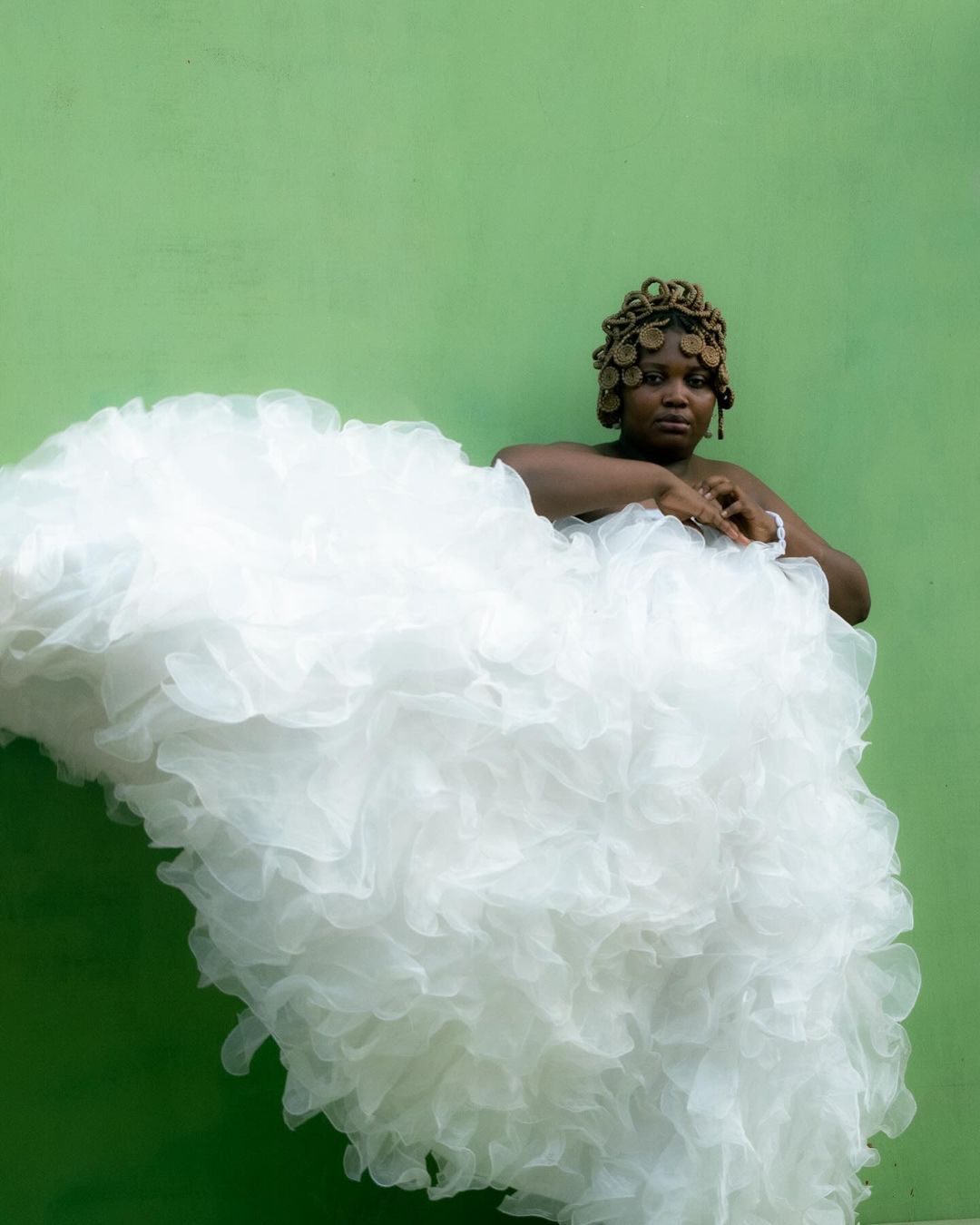 Woman poses in ruffled white dress against bright green wall, wearing unique hair accessory. Beauty shot.