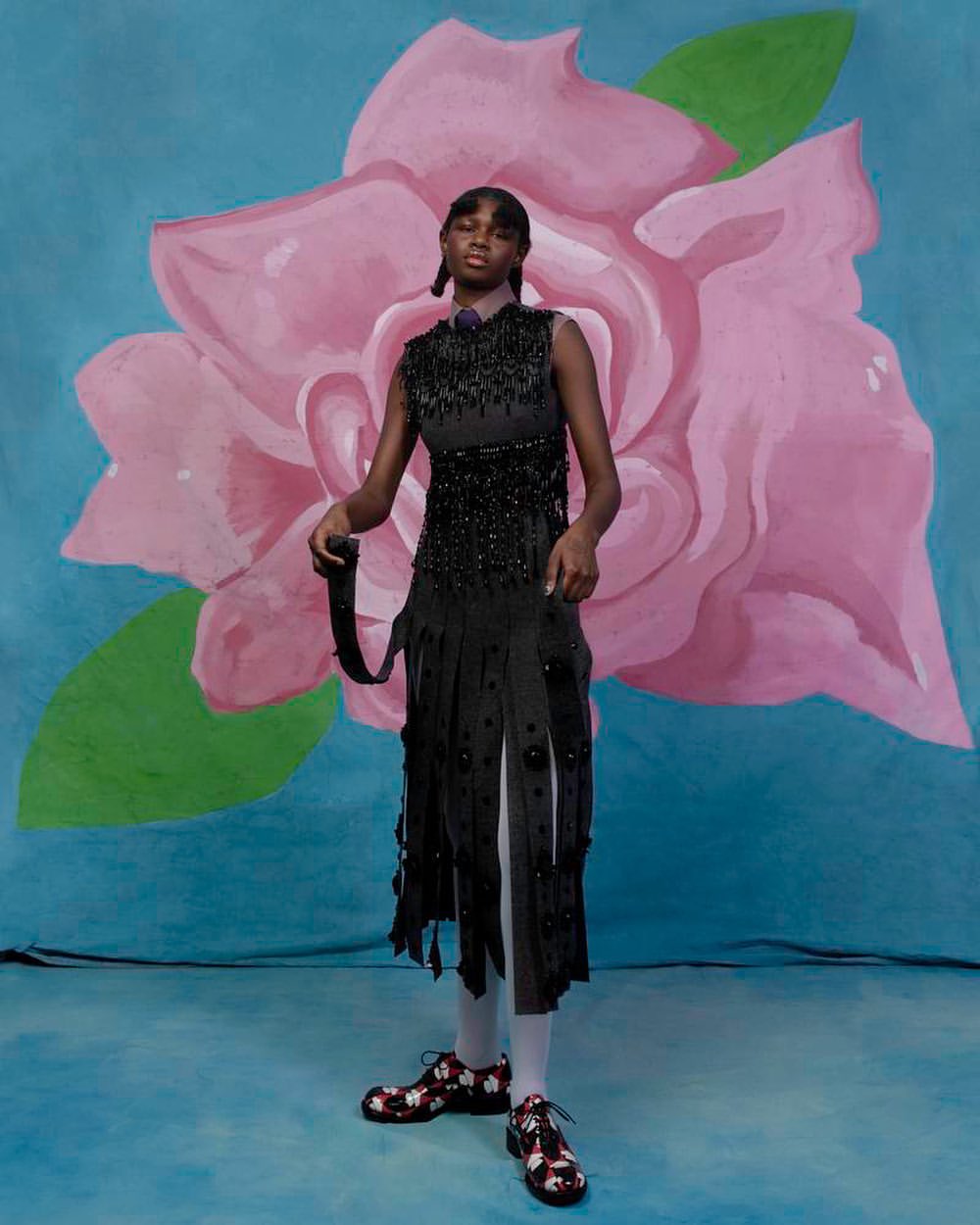A Black female model stands in a black fringed dress, against a backdrop with a large rose illustration in a fashion editorial shot.