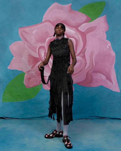 A Black female model stands in a black fringed dress, against a backdrop with a large rose illustration in a fashion editorial shot.