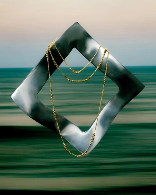 Abstract photograph featuring a gold necklace draped inside a large, silver, diamond-shaped frame with a blurred ocean and sky background.