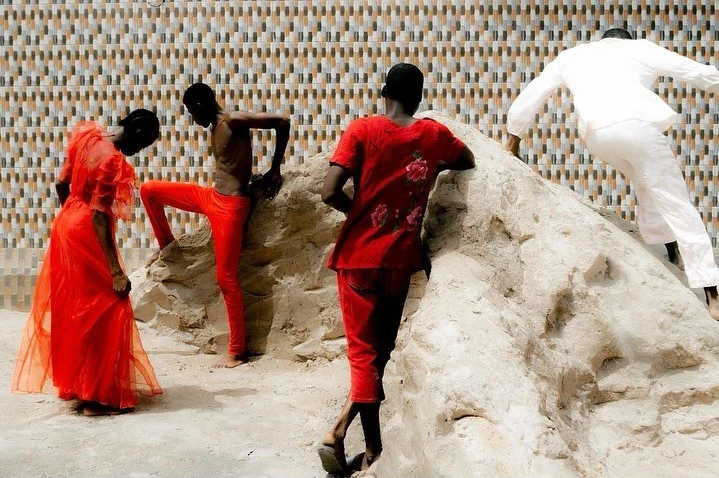 Four figures in a bright outdoor scene: one woman in a red dress, two figures in red clothing, and one figure in white atop a rock formation.