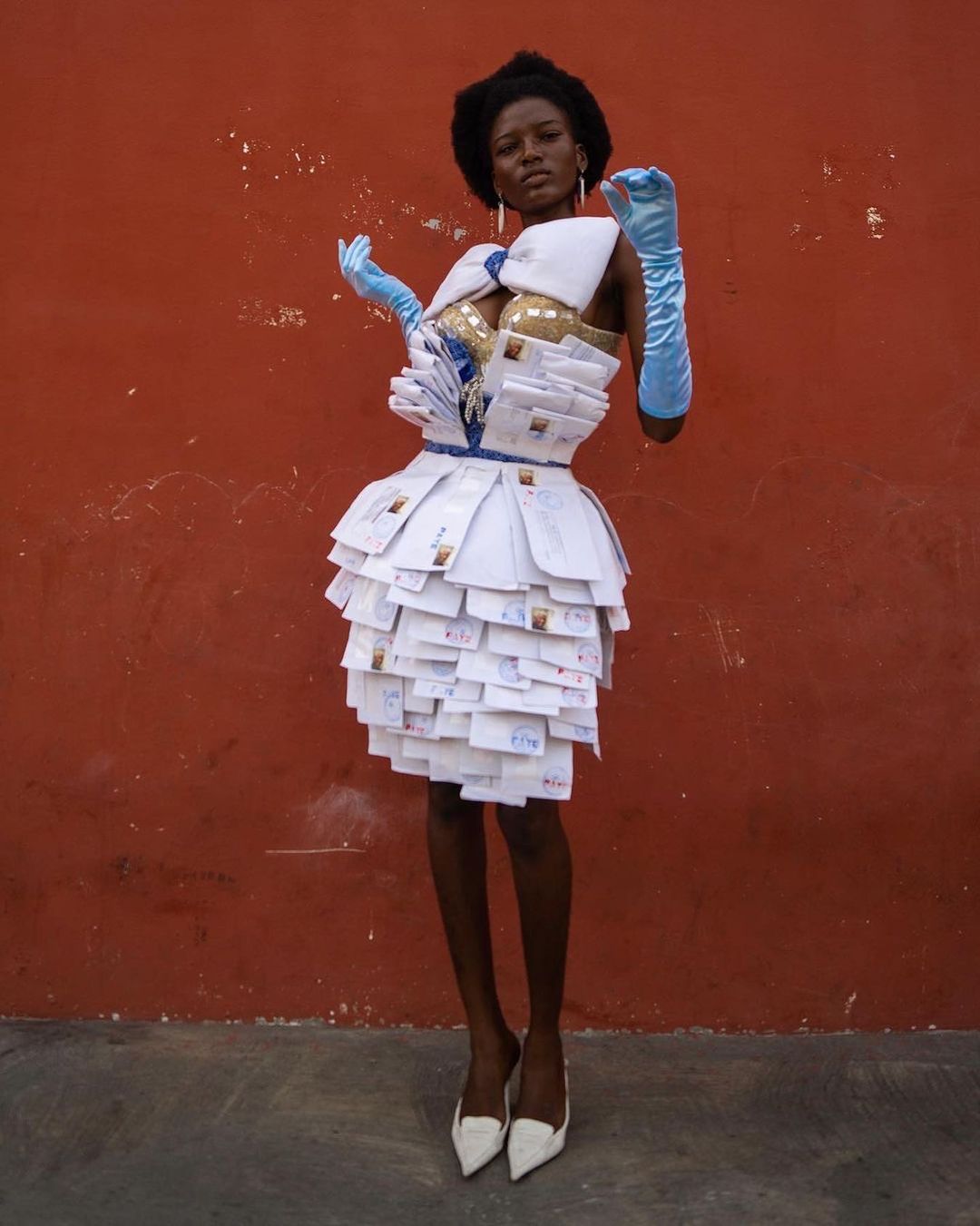 Woman poses in dress made of envelopes against red background, accessorized with blue gloves and white heels for high fashion statement.
