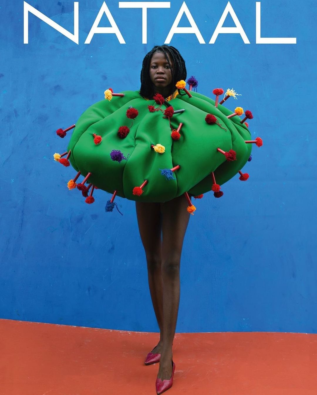 A Black woman in an avant-garde green dress with pom-poms poses against a blue wall with an orange floor, fashion, and studio lighting.