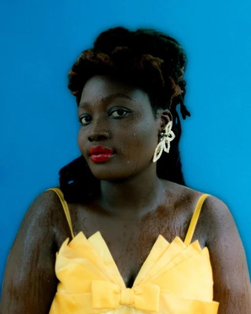 Close-up of a black adult female in a yellow dress and floral earrings against a blue background in a fashion portrait.
