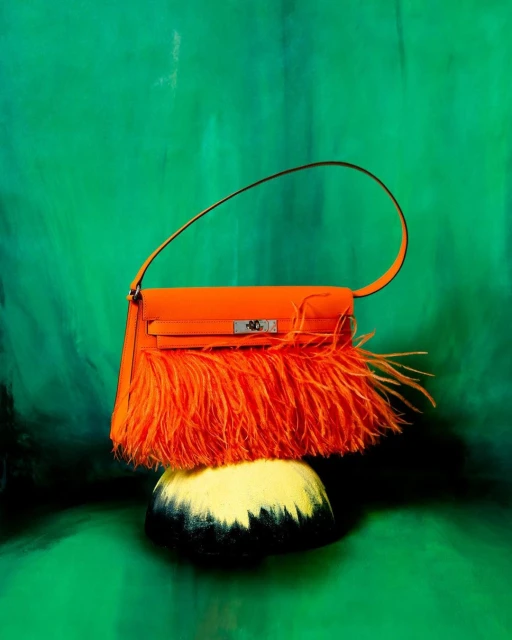 A bright orange handbag with feather-like fringe sits on a black and yellow painted base against a painterly green backdrop.