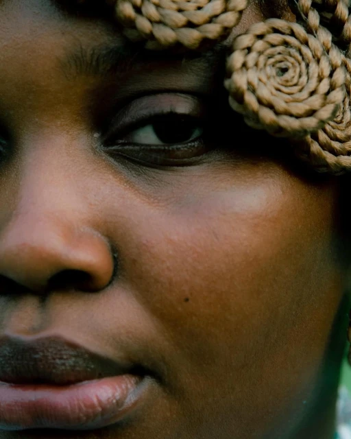 Close-up portrait of a Black woman with intricate braided spirals woven into her hair, set against a soft-focus outdoor backdrop.