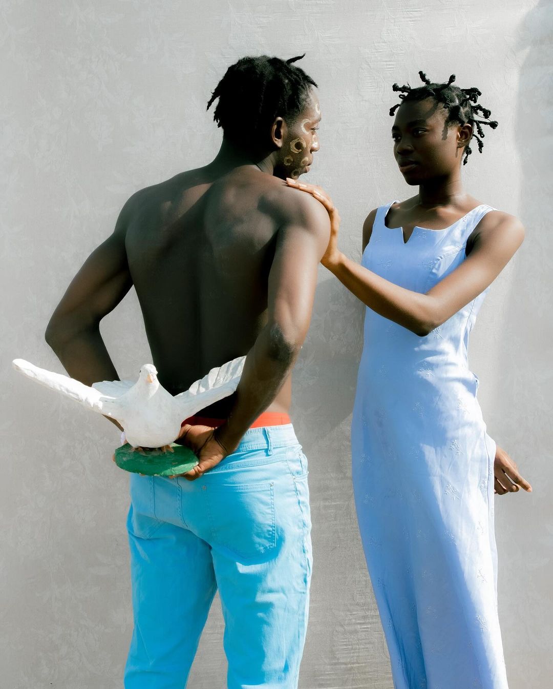 Striking portrait of an African couple in matching light blue clothing, posing against a neutral wall with a white dove at the man's back.