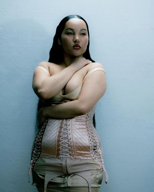 A nude woman with fair skin poses in a peach-colored corset, bra, and garter belt against a light gray background, looking away.