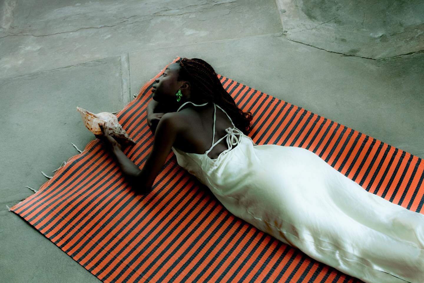 A dark-skinned woman with braids lies on a striped rug, holding a conch shell while wearing a silky white dress indoors.