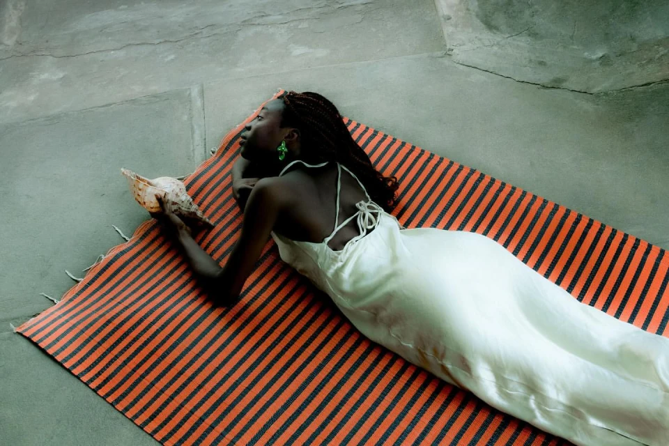 A dark-skinned woman with braids lies on a striped rug, holding a conch shell while wearing a silky white dress indoors.