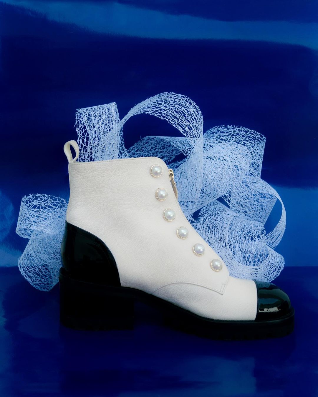 Elegant ankle boot with a color-blocked design, featuring cream leather, black toe cap and heel, pearl embellishments, and a sleek, shiny blue background.