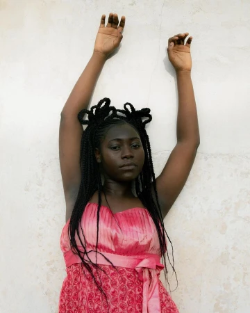 Black woman in a pink dress with braided hair posed against a textured wall with arms raised
