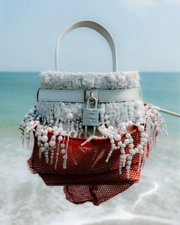 Fashionable white handbag with beaded accents and a lock detail is displayed against a blurred background of blue sea and sky.