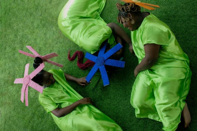 Three dark-skinned women in shiny green dresses lay on green grass, wearing colorful hair accessories. Shot from overhead.