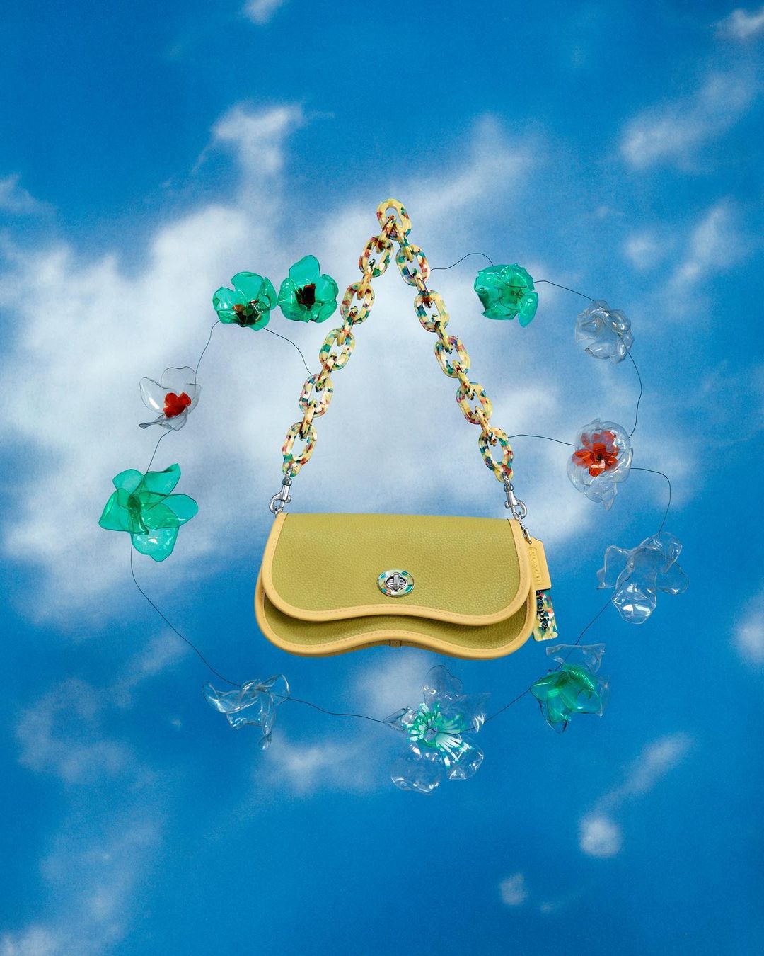 Yellow purse with a patterned chain strap, displayed with clear and teal plastic floral decorations against a blue sky backdrop.