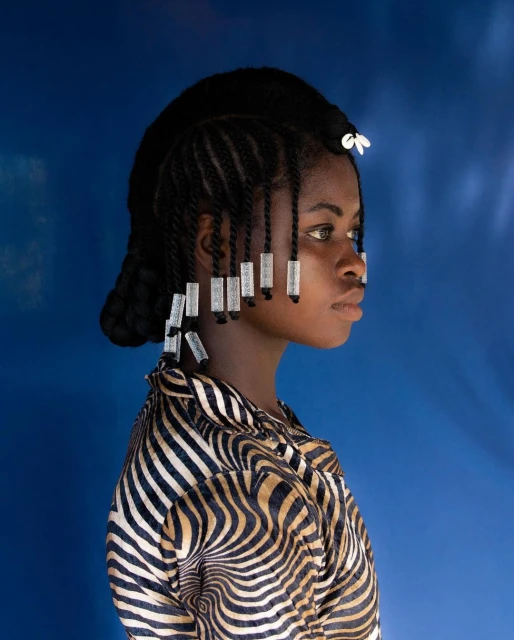 Profile portrait of a young Black female with braided hair and shell decorations against a blue studio backdrop, wearing a patterned shirt.