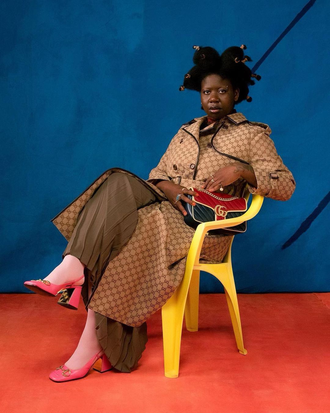 An adult woman sits on a yellow plastic chair, wearing a tan Gucci-print trench coat, pleated skirt, pink shoes, and holding a Gucci purse.