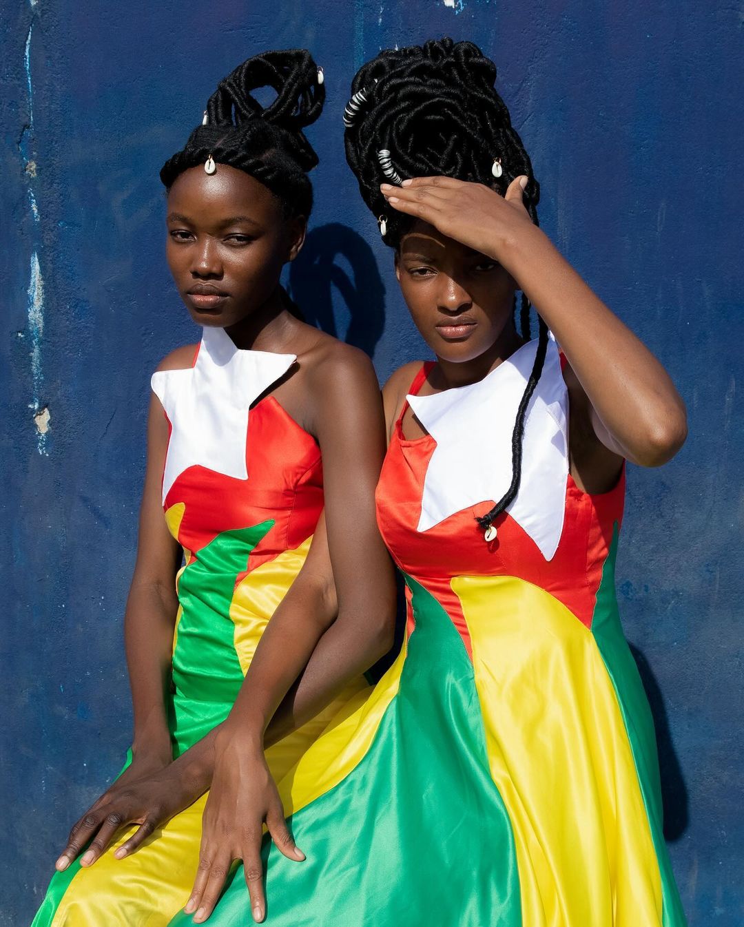 Two women in vibrant gowns pose against a blue wall. The gowns feature red, yellow, green, and white sections in a striking design.