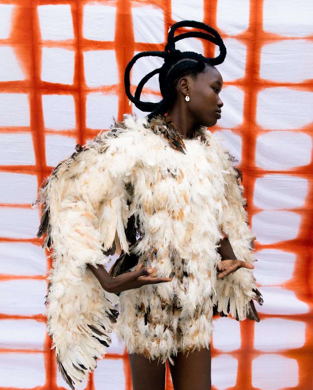 Woman in a feathered jacket poses against a white and orange grid backdrop, showcasing avant-garde fashion and unique hair styling.