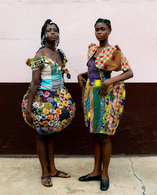 Two African women showcasing vibrant, patterned dresses with unique silhouettes against a neutral wall in natural lighting. Fashion editorial.
