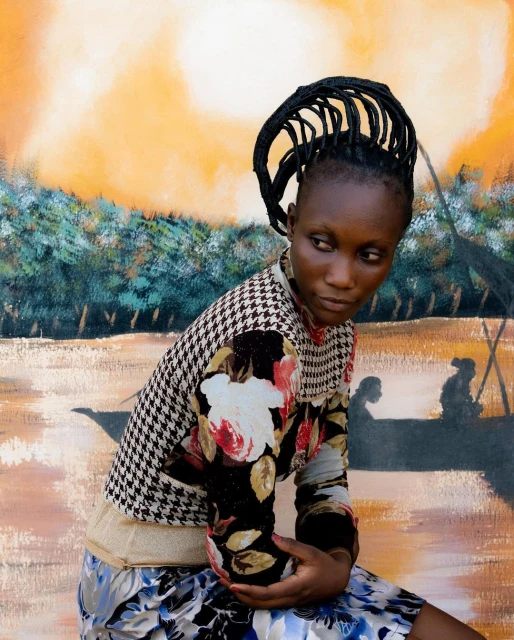 Young African woman in patterned top poses in front of a vibrant painted backdrop featuring trees and figures in a boat on the water.