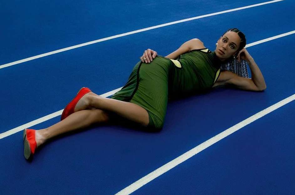 Athletic woman in green dress and red flats poses on a blue running track with white lines, lying on her side looking at the camera.