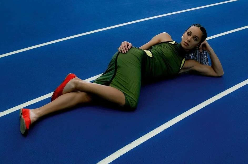 Athletic woman in green dress and red flats poses on a blue running track with white lines, lying on her side looking at the camera.