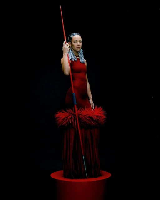 A woman poses in a red dress, holding a red spear, in a studio with a black background. She stands on a short red column.